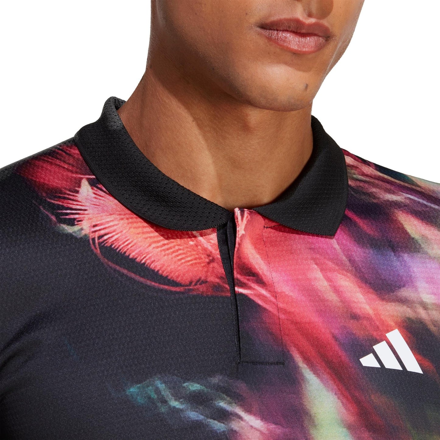 A person wearing the adidas Melbourne Men's Freelift Tennis Polo Shirt in black and multi-color patterns of red, pink, and black. The polo features a black collar and is adorned with a white Adidas logo on the chest. This eye-catching design embodies FreeLift technology as the image highlights the top half of the shirt along with part of the individual's face.