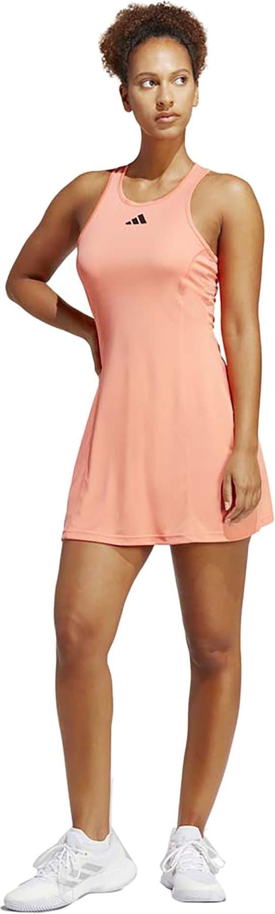 A person wearing a sleeveless adidas Women's Club Tennis Dress in coral, crafted with Primegreen fabric, and white sneakers, stands with one hand on their hip. Their curly hair is tied back as they gaze to the side, showcasing both style and the comfort provided by AEROREADY technology.
