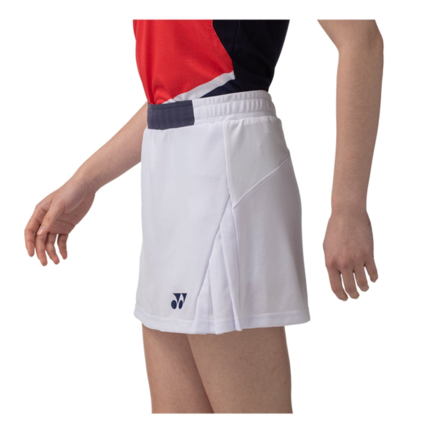 A person is dressed in a Yonex 26094 Women's Skort - White, crafted for UV reduction, with arms slightly bent. The red and black top pairs well with the white athletic skirt, displaying a subtle logo on the side. The image showcases a side profile from shoulders to knees.