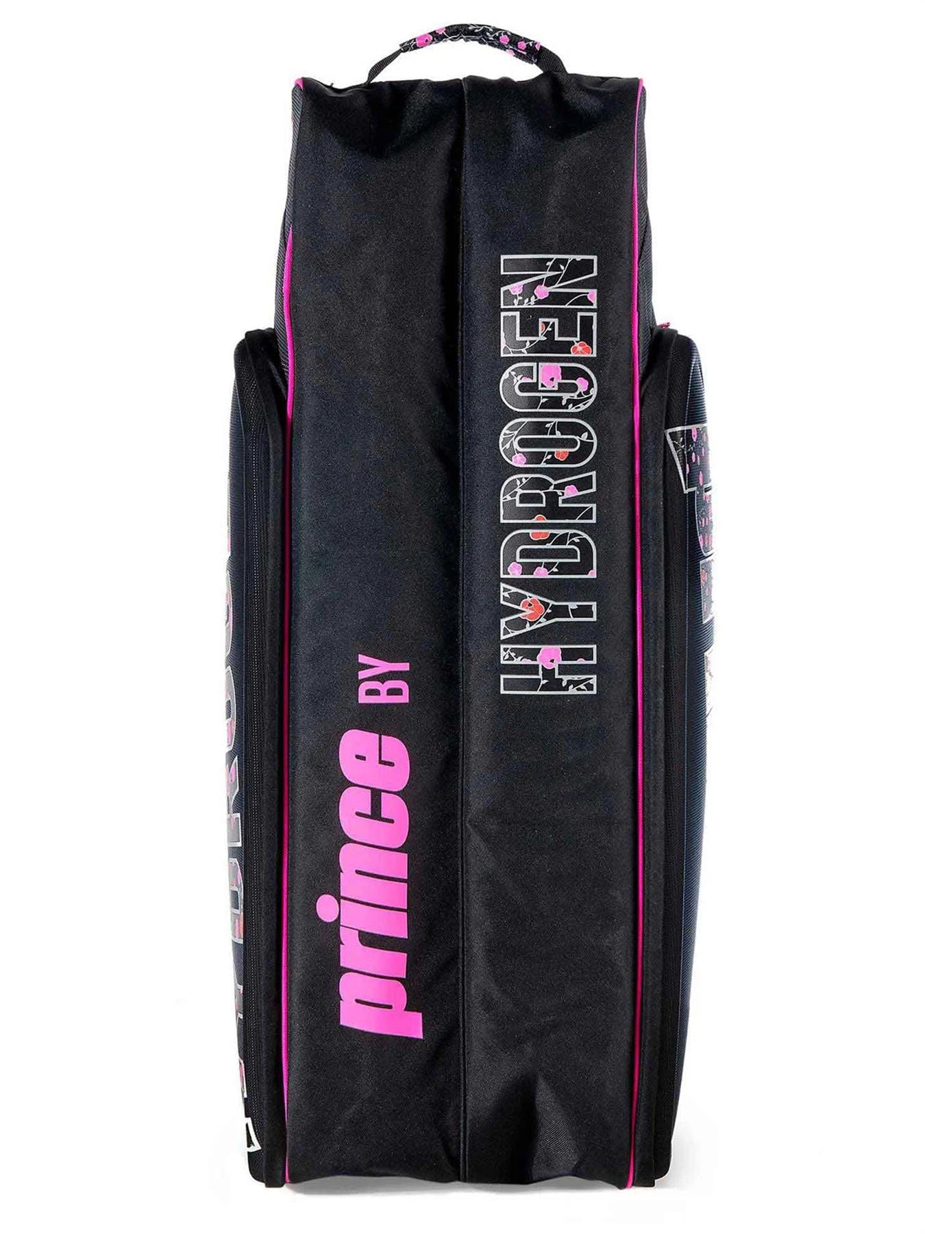 The Prince Hydrogen Lady Mary 6 Racket Tennis Bag in black with pink accents offers a stylish and functional design. It can accommodate up to six rackets, features the words "Prince by" on one side, and displays "HYDROGEN" on the other with a skull design integrated into the letter "O." This bag, equipped with a top handle and elegant pink edge accents, is ideal for those who value both fashion and functionality.