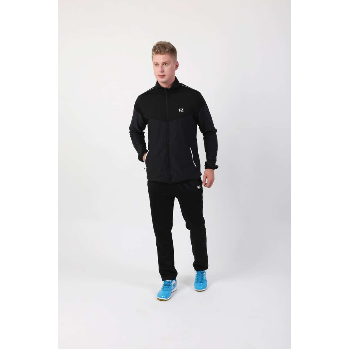A person wearing the FZ Forza Bradford Jacket in black and blue sneakers stands against a plain white background. They appear to be in motion, as if walking, perhaps ready for upcoming tournaments.