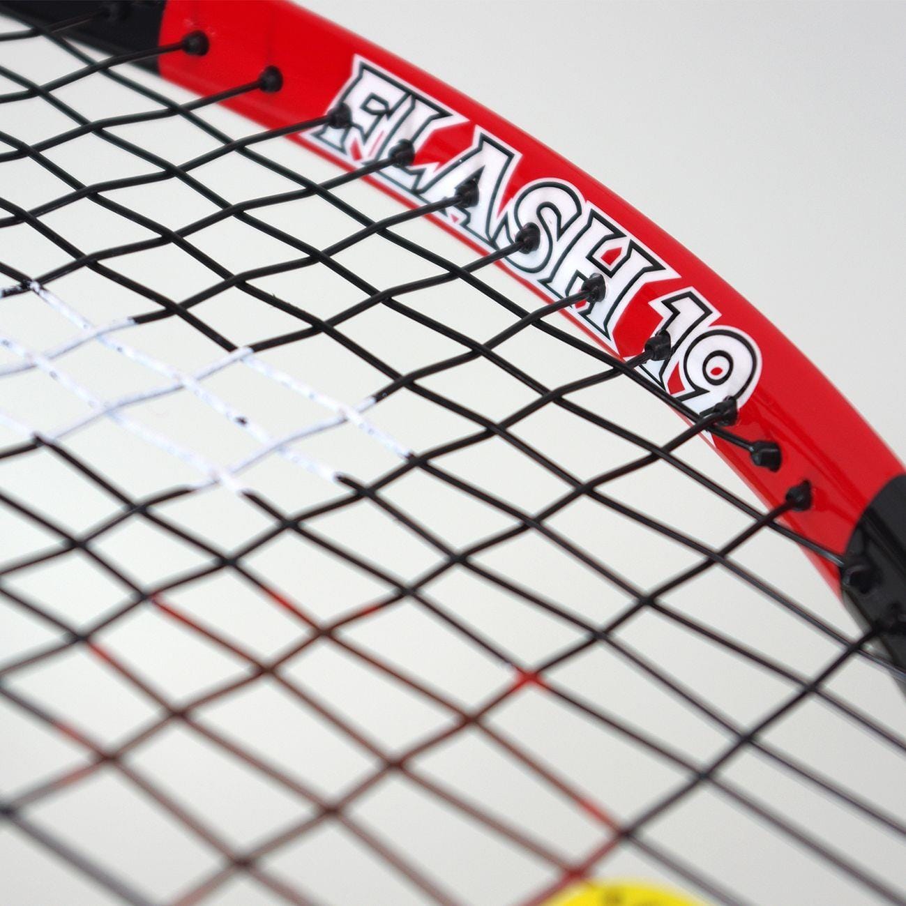 Close-up of the Karakal Flash 19 Junior Tennis Racket in black and red, featuring "FLASH 19" on its frame. Constructed from durable 7050 Alumag Alloy, the racket showcases tightly woven strings with a blurred yellow tennis ball visible underneath.