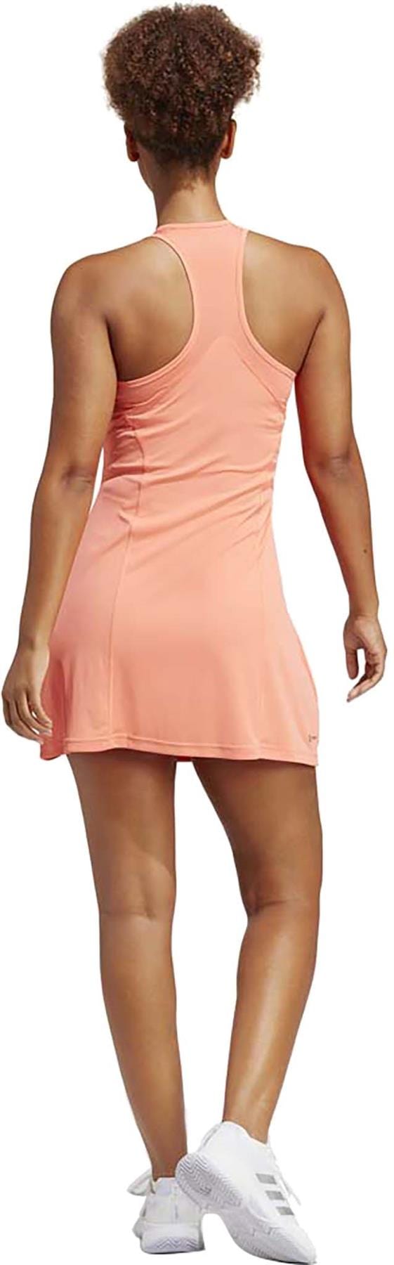 A person wearing a sleeveless coral athletic dress, similar to the adidas Women's Club Tennis Dress with AEROREADY technology, is paired with white sneakers and stands facing away. With curly hair against a pristine white background, they embody both style and sustainability reminiscent of Primegreen materials.