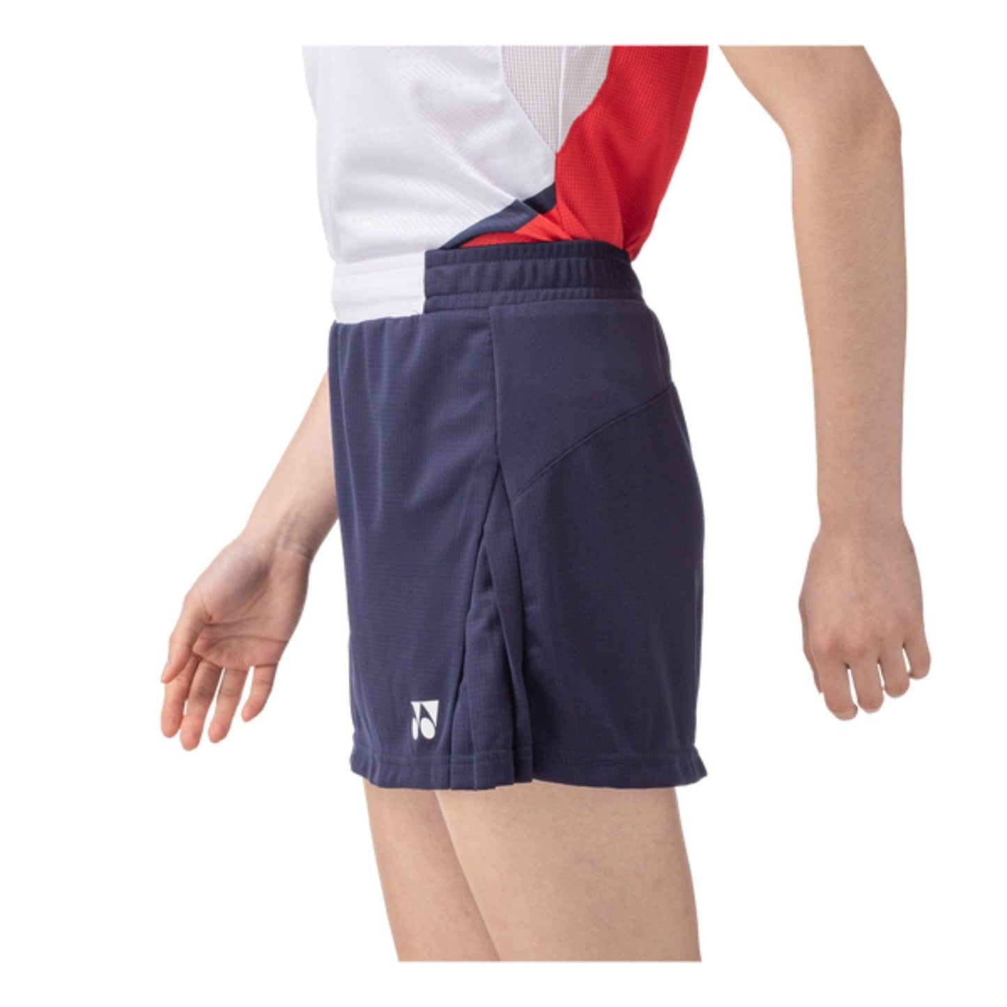A person is sporting a white and red sleeveless top combined with the Yonex 26094 Women's Skort in navy blue, highlighting their athletic style. The skort includes UV reduction technology and displays a discreet logo while they stand sideways, emphasizing the outfit.