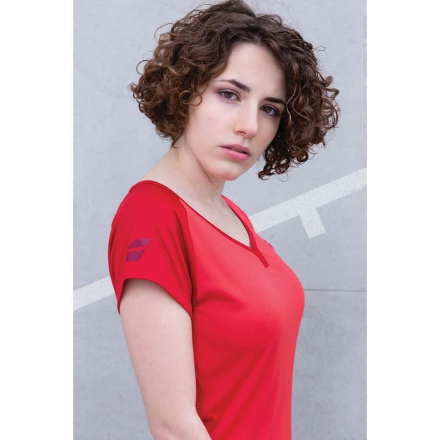 A person with curly hair wearing a red Babolat Play Womens Cap Sleeve Tennis Top, crafted from FiberDry fabric, stands against a light gray background, looking at the camera with a neutral expression.