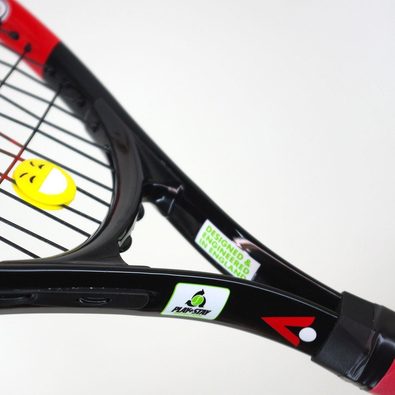 A close-up of the Karakal Flash 19 Junior Tennis Racket - Black / Red highlights its black frame with yellow strings, complemented by a smiling emoji vibration dampener. The frame is crafted from 7050 Alumag Alloy and adorned with various logos and vibrant designs, making it visually striking.