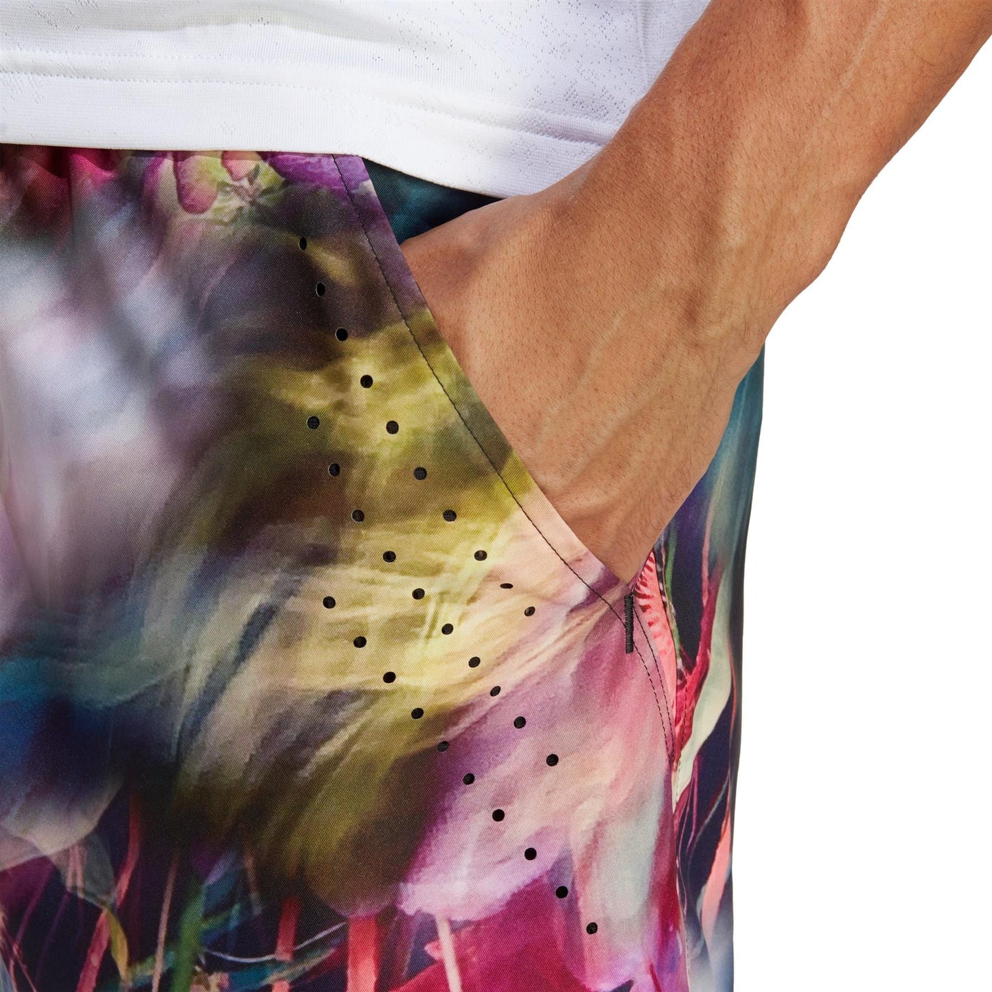 A close-up of a hand tucked into the pocket of vibrant, floral-patterned shorts, crafted from recycled materials. The design features multicolored abstract flowers with subtle black perforations. The person pairs this look with a crisp white shirt, wearing the adidas Melbourne Men's Ergo Graphic Tennis Shorts in Black/Multi by adidas.