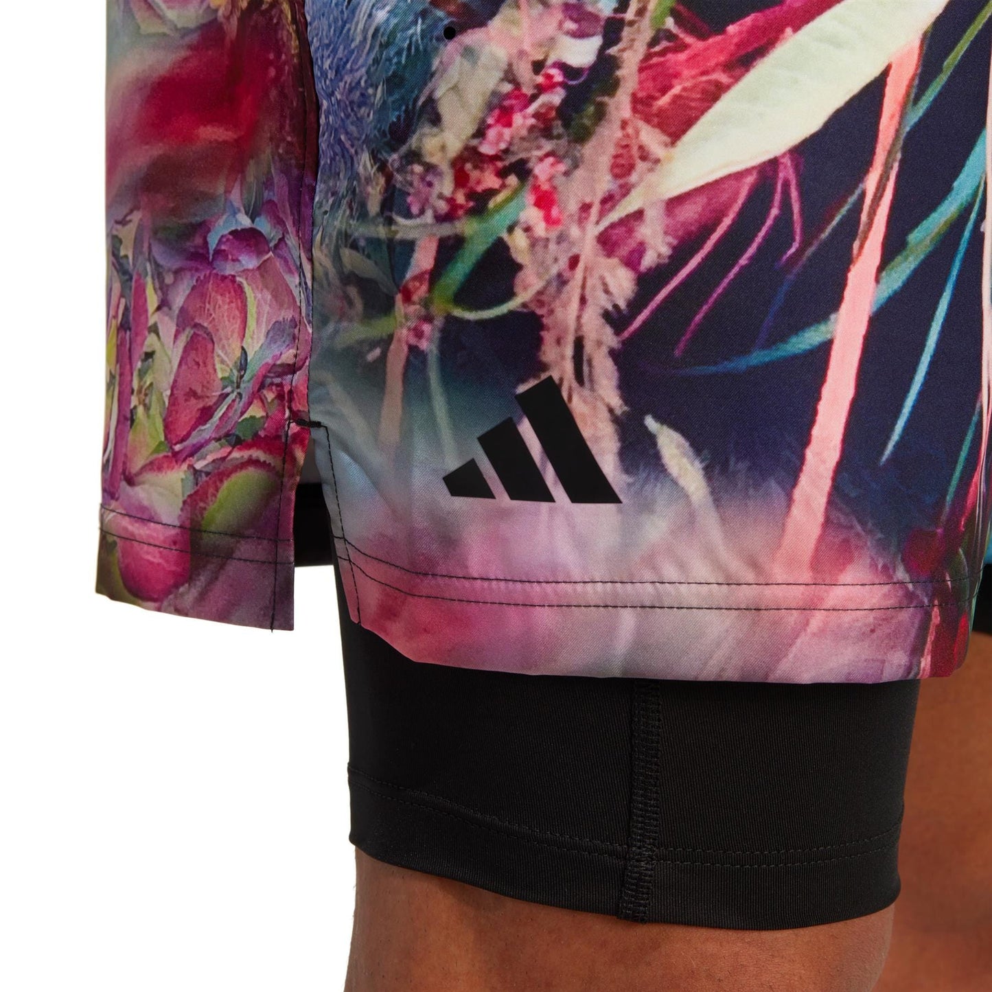 Close-up of a person's leg wearing the adidas Melbourne Men's Ergo Graphic Tennis Shorts in Black/Multi, featuring vibrant floral patterns. These shorts, crafted from recycled materials, display a small black logo with three stripes. Beneath the shorts, black leggings are visible.