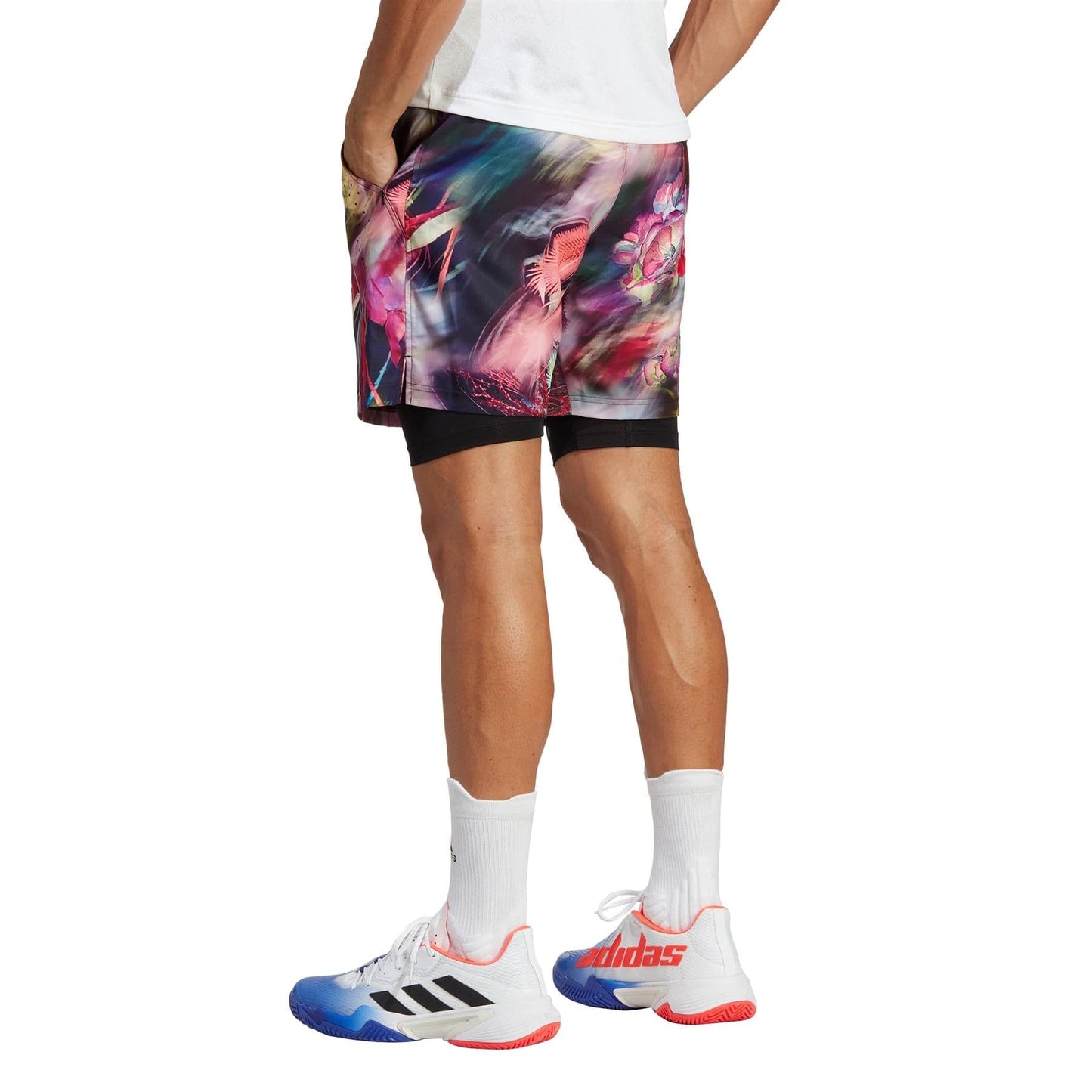 A person wearing vibrant adidas Melbourne Men's Ergo Graphic Tennis Shorts in Black/Multi, paired with a white shirt and Adidas sneakers featuring blue and red accents. The image focuses on the lower body, showing their hands in pockets and white socks on a light gray surface.