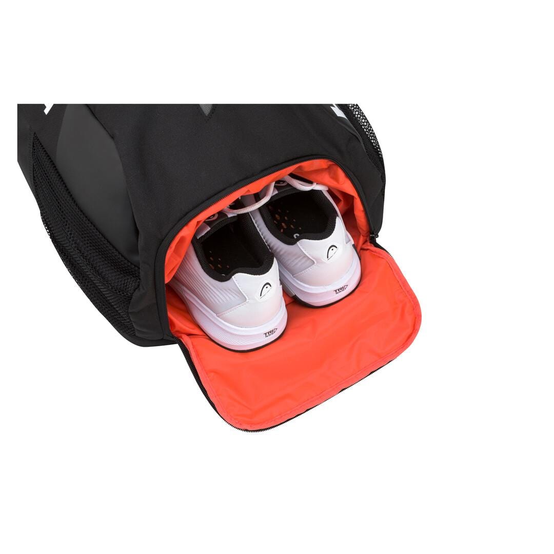 The HEAD Tour Team Tennis Backpack in Black/Orange sits partially unzipped, showcasing a pair of white tennis shoes nestled in its designated shoe compartment. The bright orange interior offers a striking contrast to the sleek exterior and the white shoes.