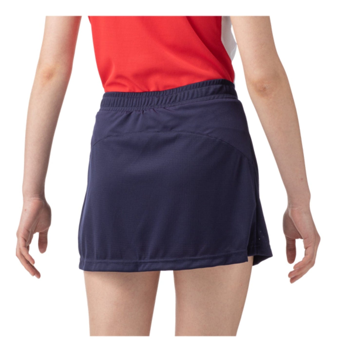 A person is wearing a red top along with the Yonex 26094 Womens Skort in navy blue. They face away from the camera with their arms slightly lifted, presenting a chic and functional appearance intended for UV protection against the simple white background.
