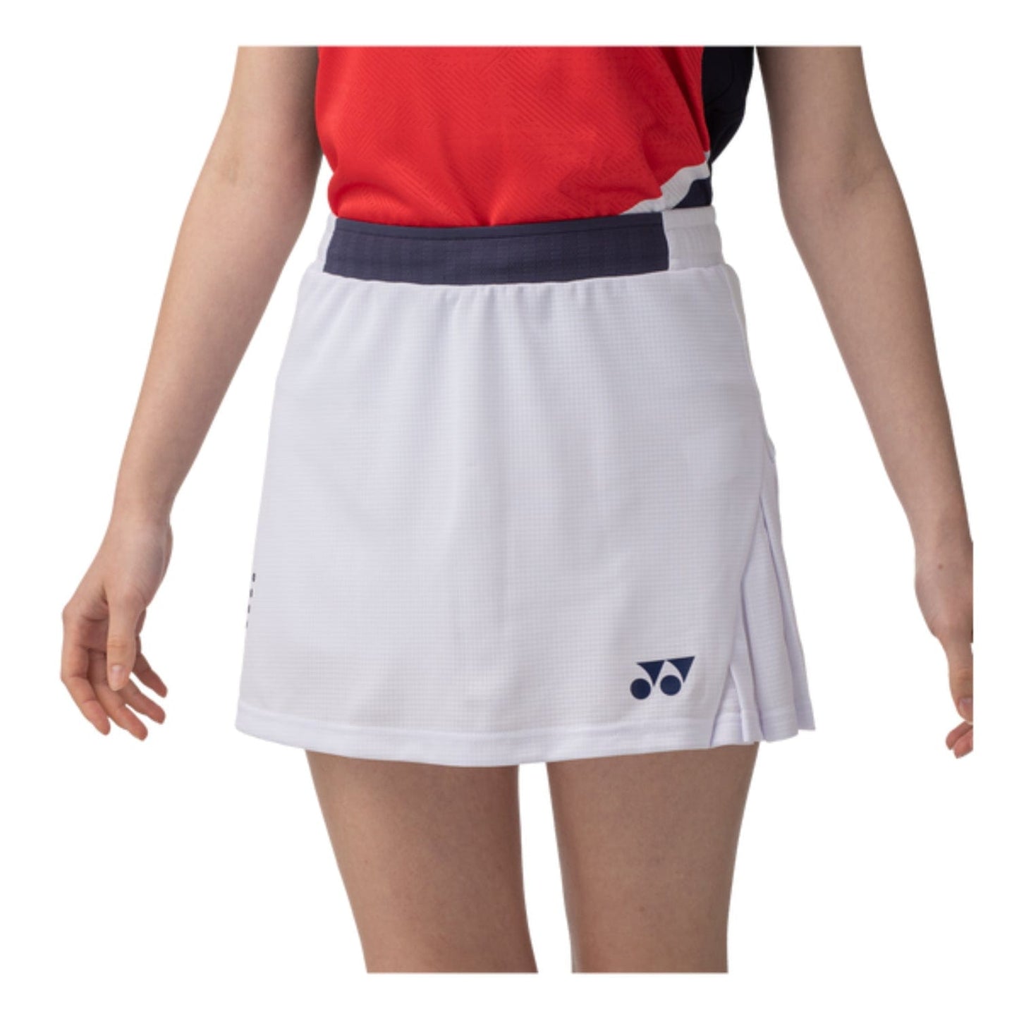 A person is wearing a red sports top and a Yonex 26094 Womens Skort in white, featuring the brand logo near the hem. Their arms are relaxed by their sides, and the photo is cropped at the shoulders and thighs.