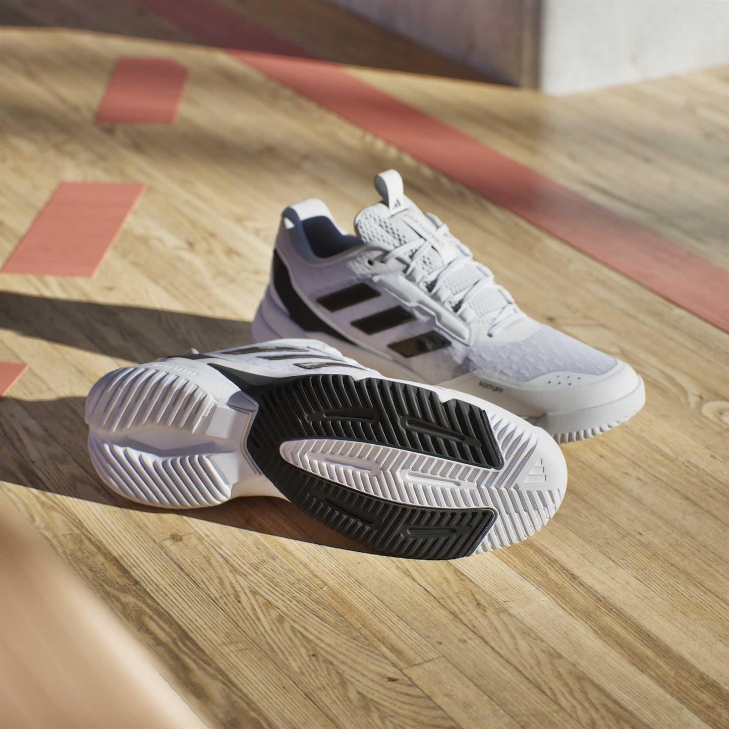 A pair of adidas Crazyflight 6 Men's Indoor Tennis Shoes in white are on a wooden gym floor; one stands upright and the other lies on its side, showing the black and white sole.
