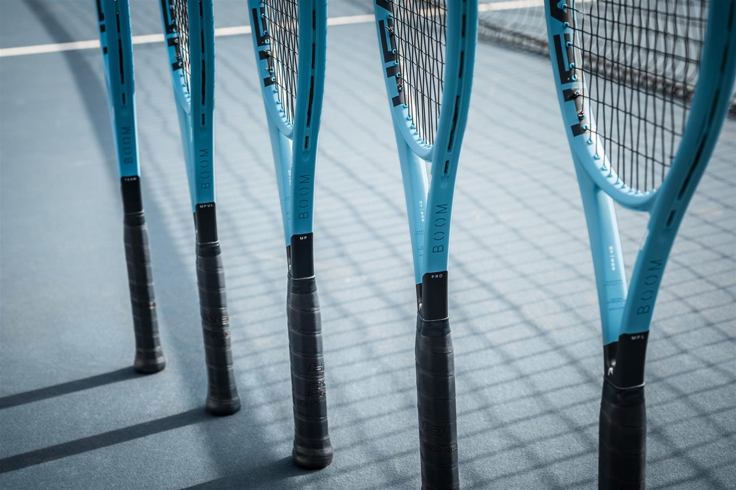 Five HEAD Boom MP L 2026 Tennis Rackets stand upright on a blue tennis court in the sunlight, their black handles casting shadows from the net, showcasing HEAD’s promise of effortless power with every swing.