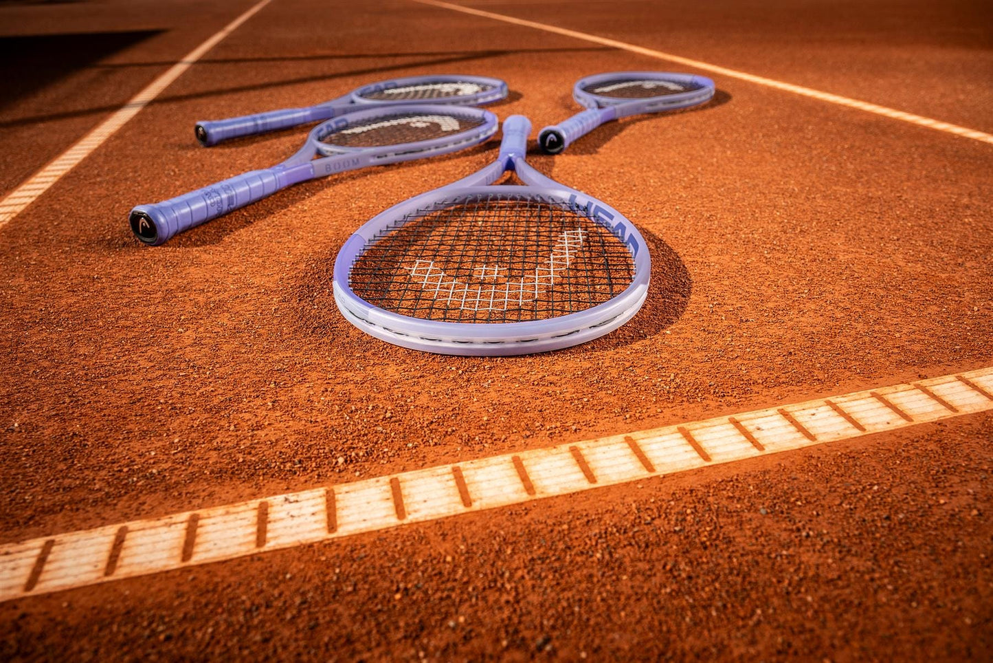 Four tennis rackets rest on a clay court under bright sunlight, with the HEAD Boom Team Alternate 2026 Tennis Racket by HEAD—ideal for beginner to intermediate players—prominently positioned near the court lines.