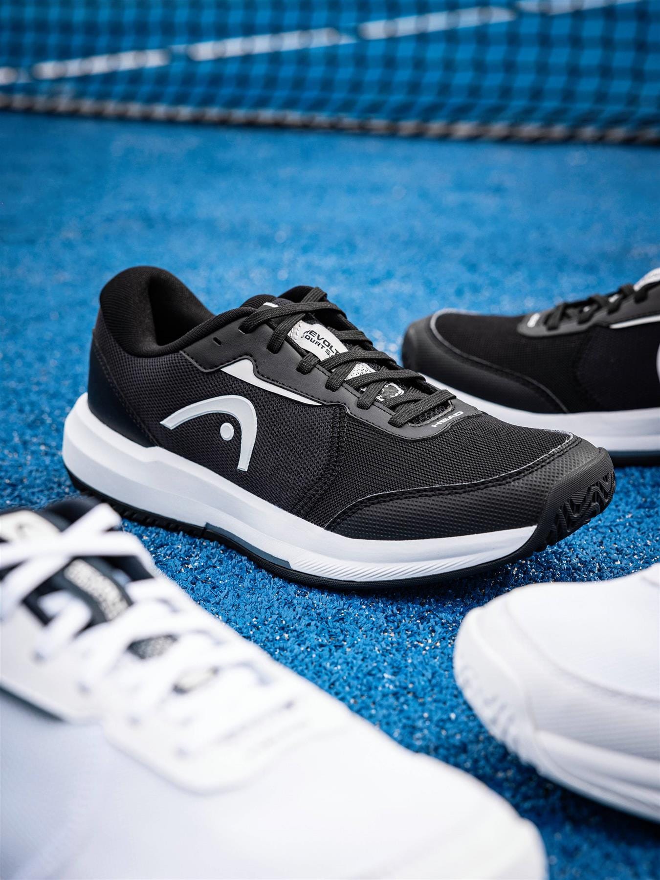 The HEAD Revolt Court 5.0 Men's Tennis Shoes in Black/White appear on a blue court, with the HEAD logo visible, white shoes nearby, and a net in the background—showcasing stylish comfort from HEAD.