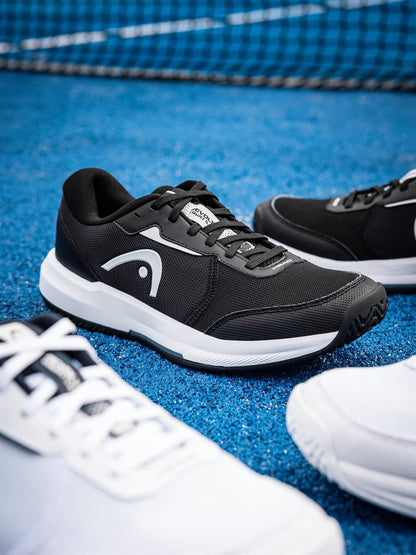 The HEAD Revolt Court 5.0 Men's Tennis Shoes in Black/White appear on a blue court, with the HEAD logo visible, white shoes nearby, and a net in the background—showcasing stylish comfort from HEAD.