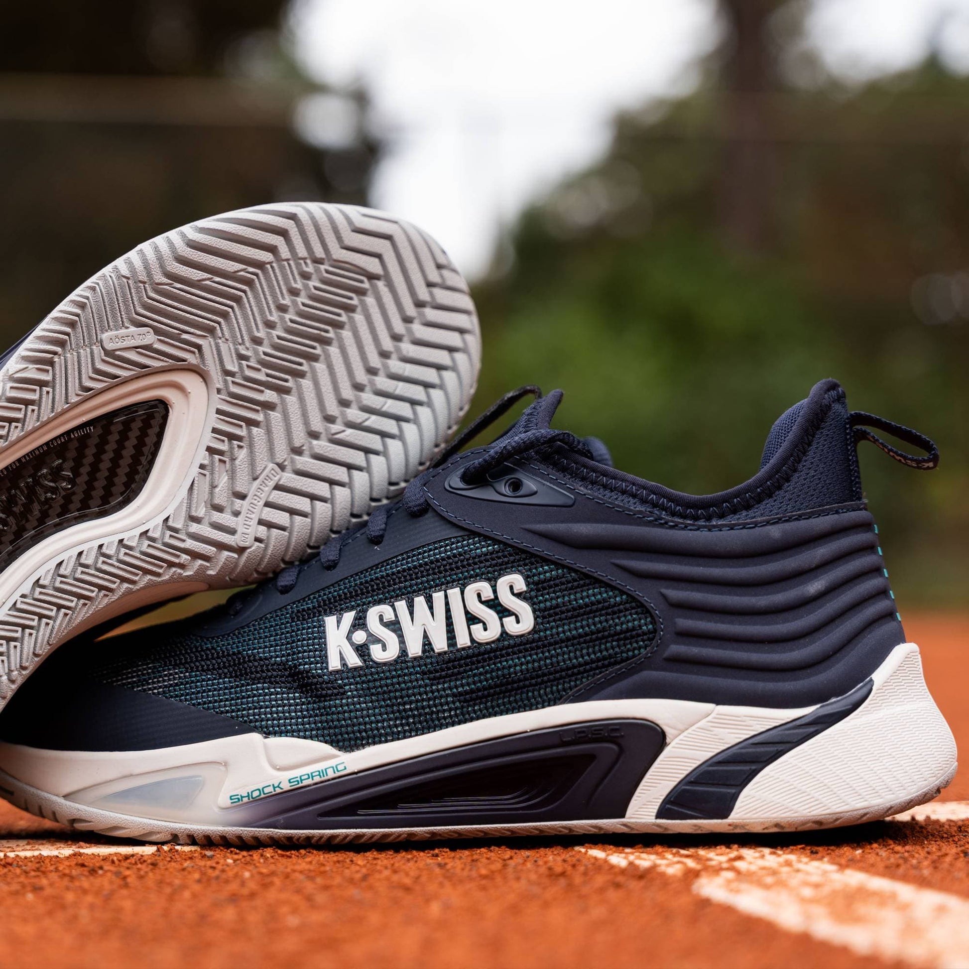 A pair of K-Swiss Hypercourt Pinnacle Men's Tennis Shoes in Carbon, White, and Baltic is showcased on a clay tennis court, with one shoe upright and the other angled to highlight its sole, tread pattern, and the advanced carbon fibre plate engineered by K-Swiss for enhanced performance.