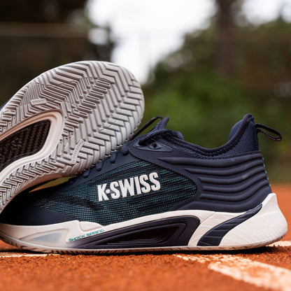A pair of K-Swiss Hypercourt Pinnacle Men's Tennis Shoes in Carbon, White, and Baltic is showcased on a clay tennis court, with one shoe upright and the other angled to highlight its sole, tread pattern, and the advanced carbon fibre plate engineered by K-Swiss for enhanced performance.