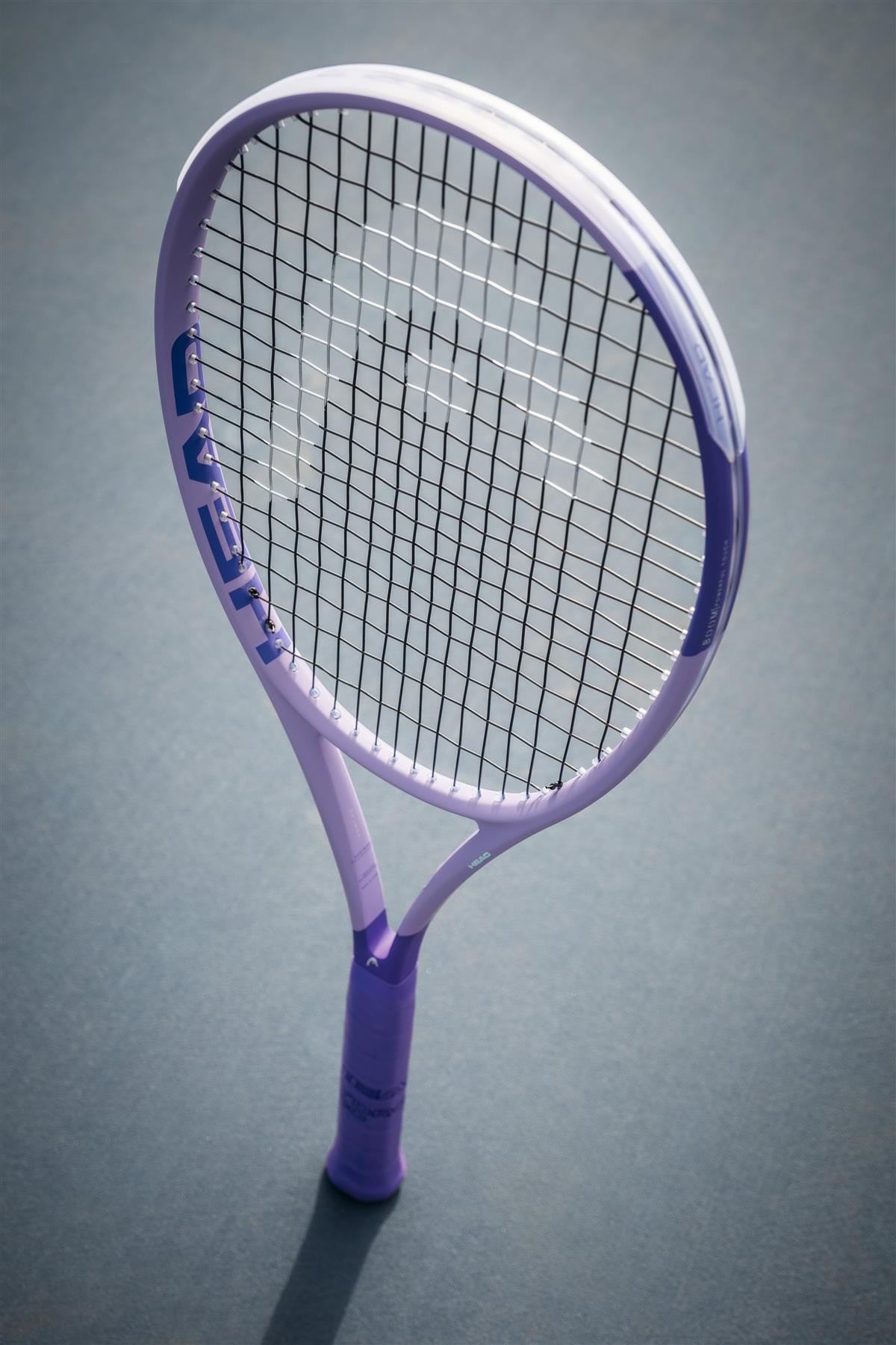 A light purple HEAD Boom MP Alternate 2026 Tennis Racket with black strings stands upright on a blue surface, casting a faint shadow—ready to deliver explosive power on the court.