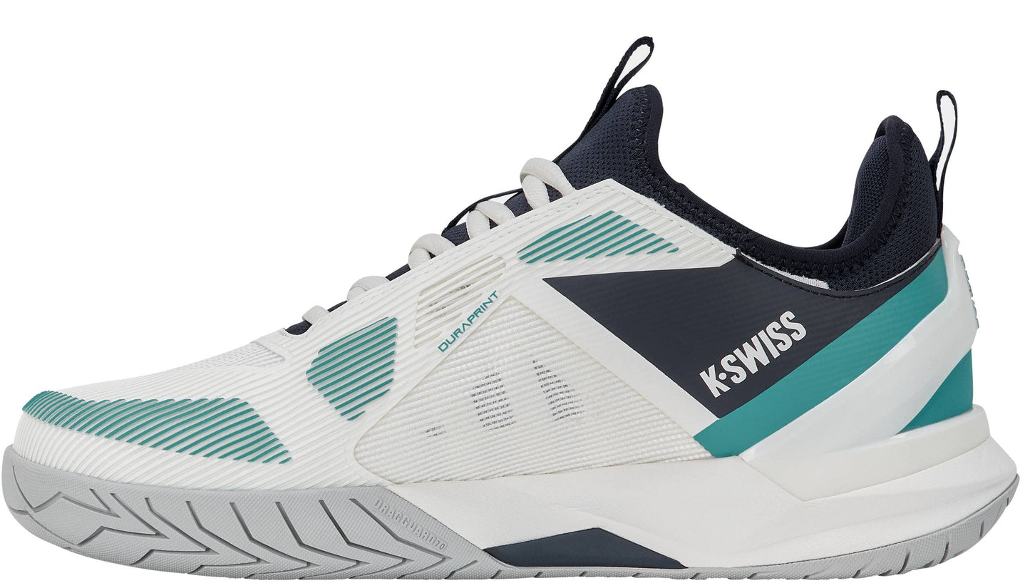 The K-Swiss K-Frame Speed Rublo Men's Tennis Shoes in White, Carbon, and Baltic feature athletic styling with teal and navy accents, a textured sole, and prominent "K-Swiss" branding on the side. The design includes mesh panels for breathability, plus a black tongue and collar. These shoes incorporate the signature K-Frame support technology—favored by pros like Andrey Rublev—for enhanced stability on the court.
