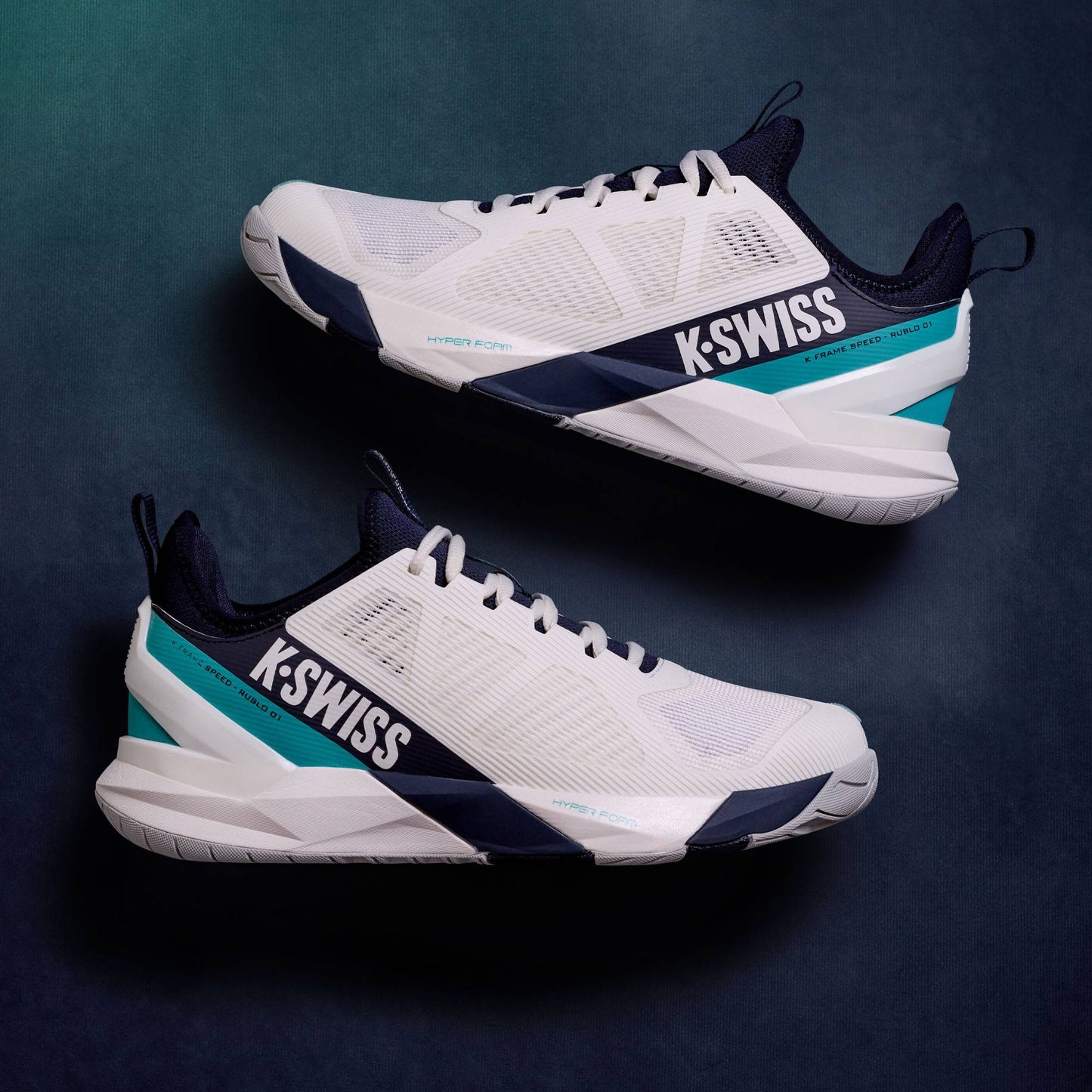 A pair of K-Swiss K-Frame Speed Rublo Mens Tennis Shoes in White, Carbon, and Baltic, showcasing the signature K-Swiss K-Frame design, are displayed side by side on a dark, textured background.