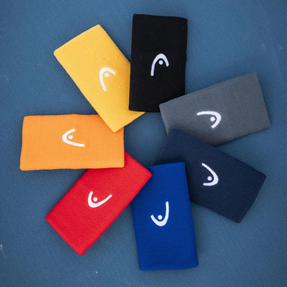 Seven rectangular HEAD 5" Tennis Wristbands—yellow, orange, red, blue, navy, gray, and black—are arranged in a circle on a blue background. Each features the white curved HEAD logo.