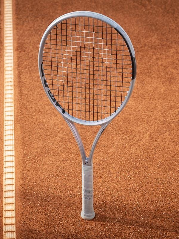 A HEAD Squared Tennis Racket by HEAD with a white grip stands upright on the clay court near the white boundary line.