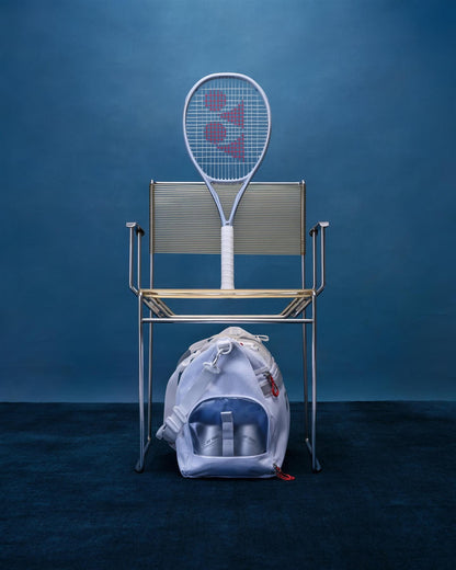 A Yonex Muse 100L Tennis Racket by Yonex rests on a sleek metal chair, with a white sports bag in front. Through the bag’s clear pocket, two bottles are visible, all set against a blue background.