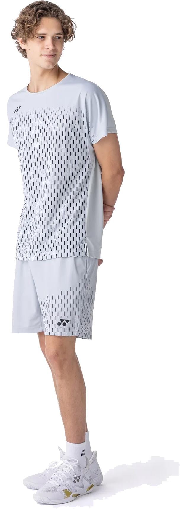 A young man stands sideways, hands behind his back, wearing a light grey sports t-shirt and Yonex 15241EX Knit Men's Tennis Shorts in Silver Grey by Yonex, featuring a black dashed pattern. He pairs the outfit with white athletic shoes.