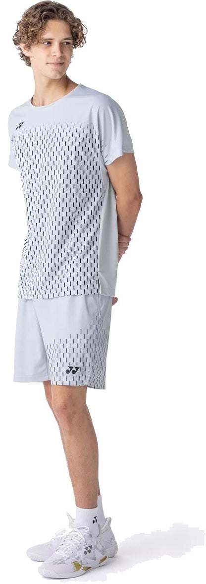 A young man stands sideways, hands behind his back, wearing a light grey sports t-shirt and Yonex 15241EX Knit Men's Tennis Shorts in Silver Grey by Yonex, featuring a black dashed pattern. He pairs the outfit with white athletic shoes.
