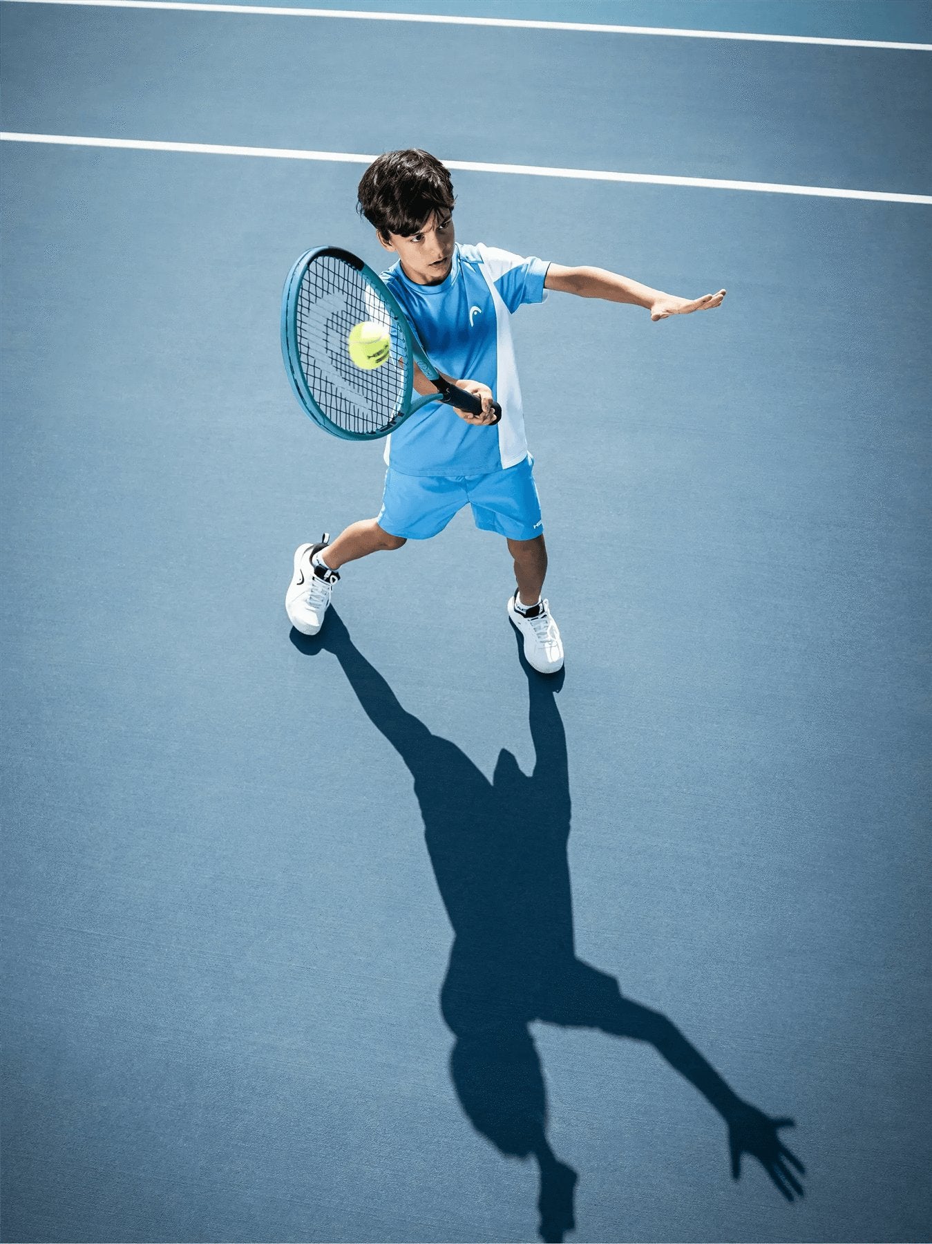 A young boy in a blue and white outfit plays tennis on a blue court, preparing to hit the ball with his HEAD Boom Jr 2026 Junior Tennis Racket. His shadow is visible on the court surface.