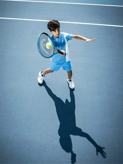 A young boy in a blue and white outfit plays tennis on a blue court, preparing to hit the ball with his HEAD Boom Jr 2026 Junior Tennis Racket. His shadow is visible on the court surface.