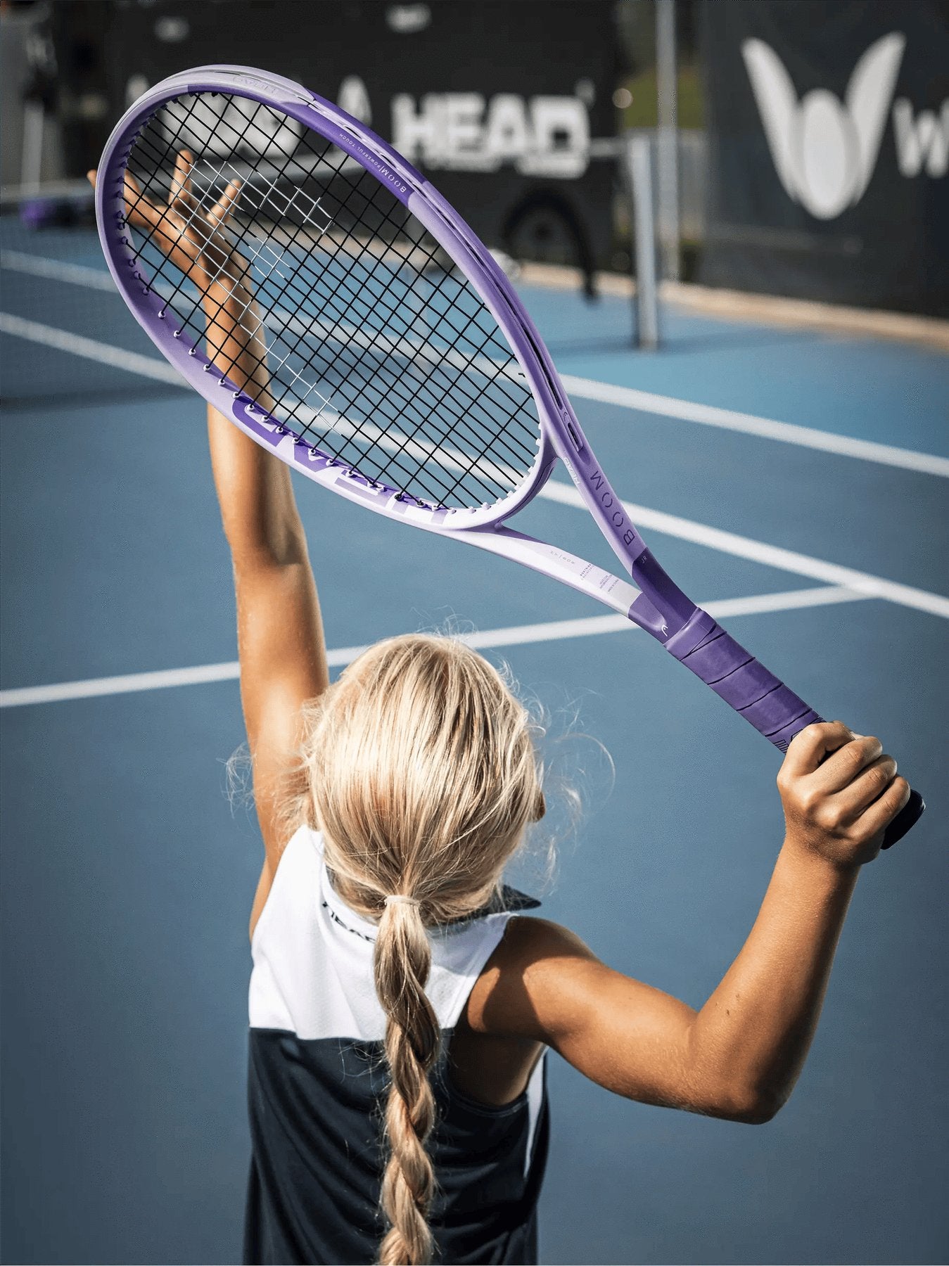 A young blonde girl with braided hair lifts her HEAD Boom Jr Alternate 2026 Junior Tennis Racket, tossing a ball up to serve on a blue outdoor tennis court.