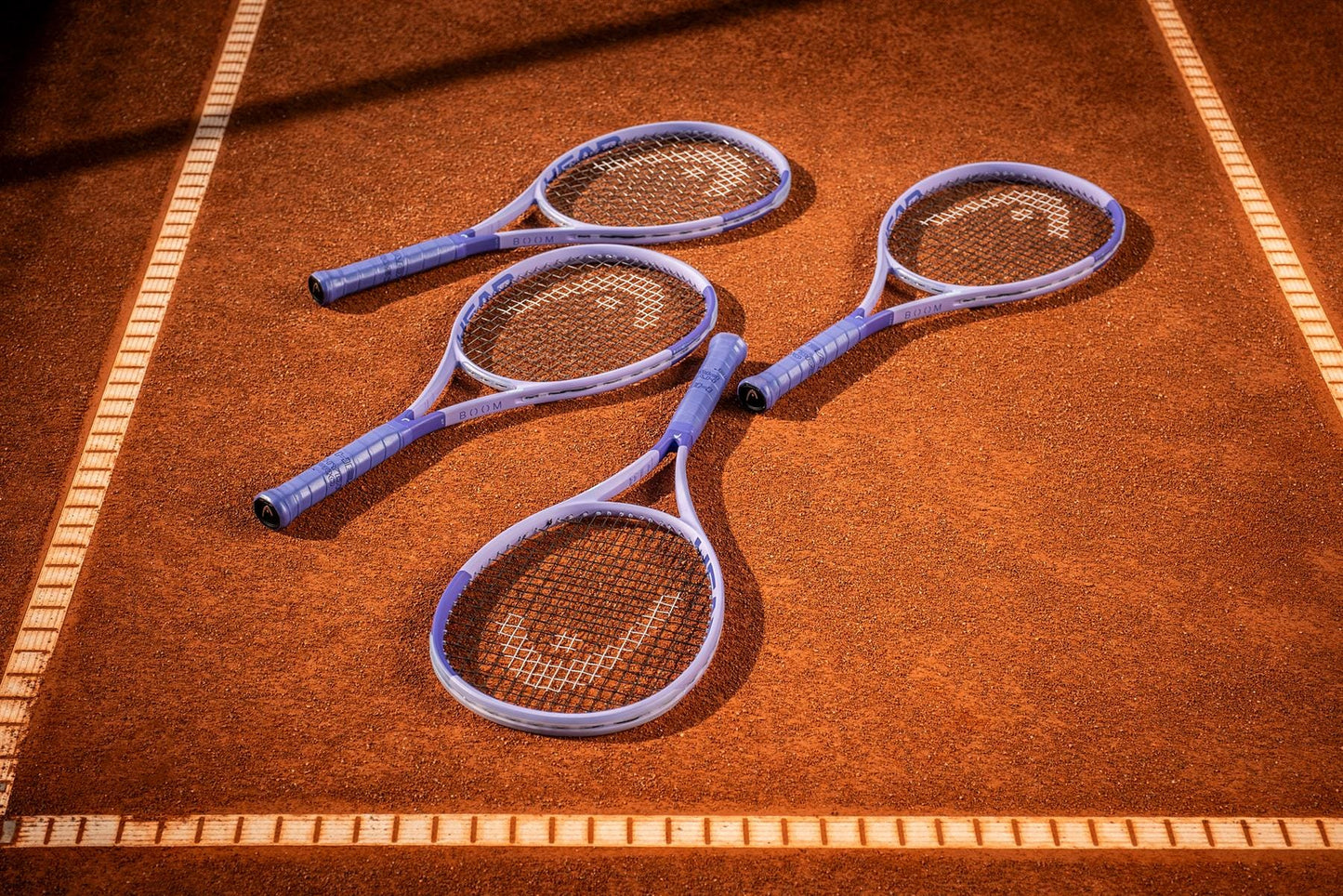 Three HEAD tennis rackets with blue handles, including the ultra-lightweight HEAD Boom MP UL Alternate 2026 Tennis Racket, lie side by side on a clay court within white boundary lines.