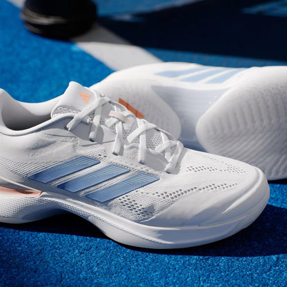 A close-up of adidas Avacourt 3 women's tennis shoes in Core White, Halo Blue, and Beam Orange rests on a bright blue tennis court with a white line visible in the background.