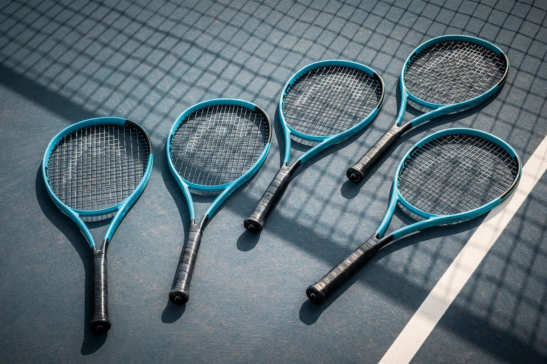 Five blue and black HEAD tennis rackets, including the HEAD Boom Team 2026 Tennis Racket, are arranged on a sunlit court near a white boundary line, their frames casting netted shadows on the surface.