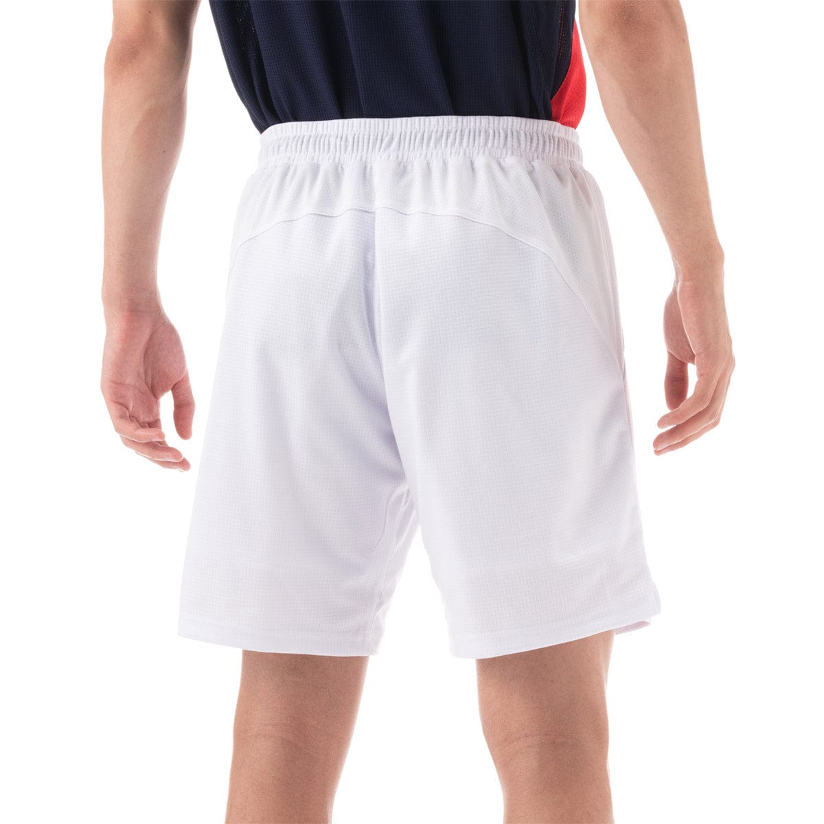 A person wearing a navy and red shirt with Yonex 15131 Mens Shorts in white stands facing away. The performance shorts from Yonex feature an elastic waistband, and their arms are relaxed at their sides against a plain white background.