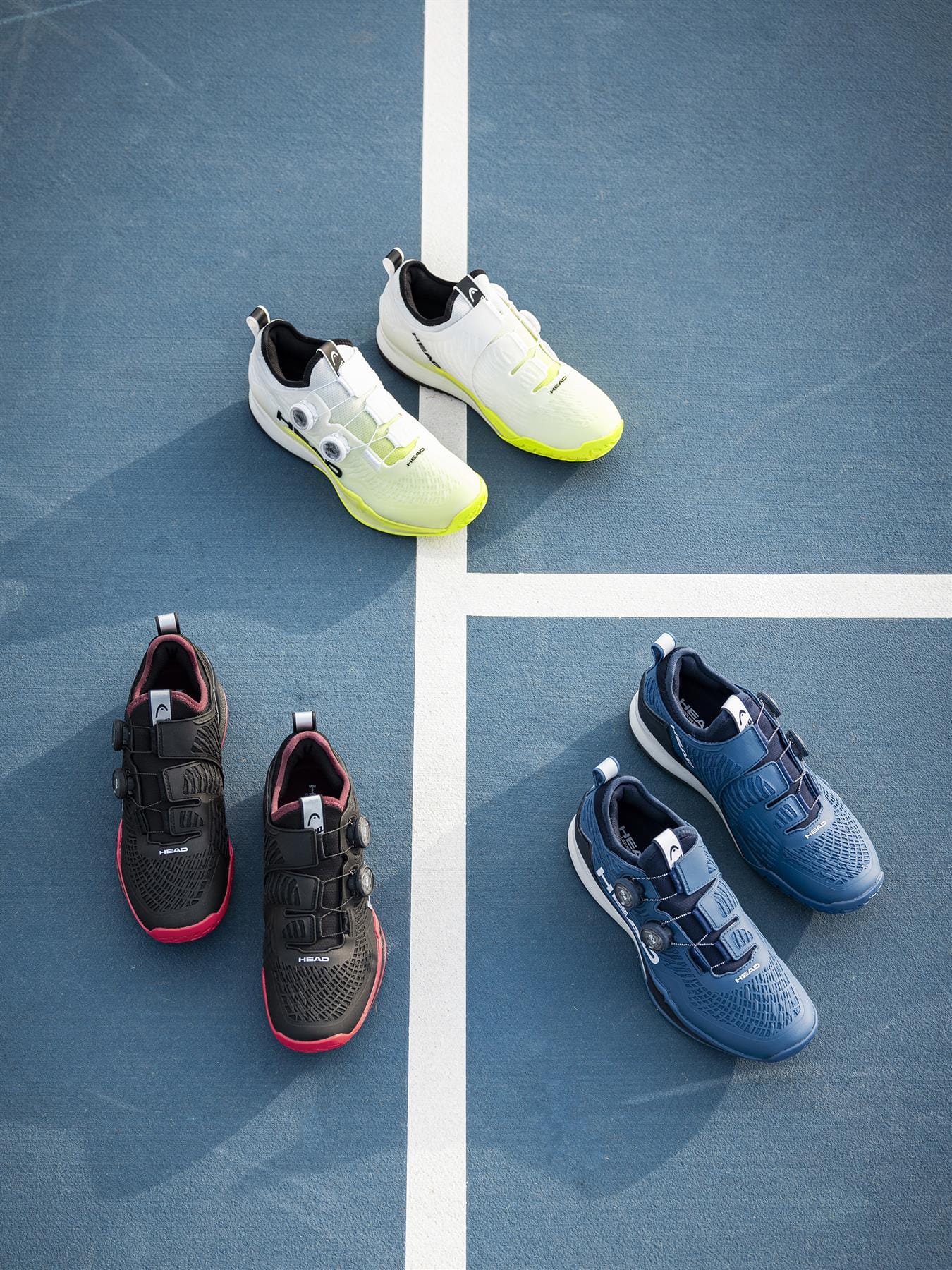 Three pairs of HEAD Endure Pro BOA Men's Tennis Shoes by HEAD in white, black with red soles, and dark blue/navy are neatly arranged on a blue sports court with white lines, highlighting their style and durability for every tennis game.