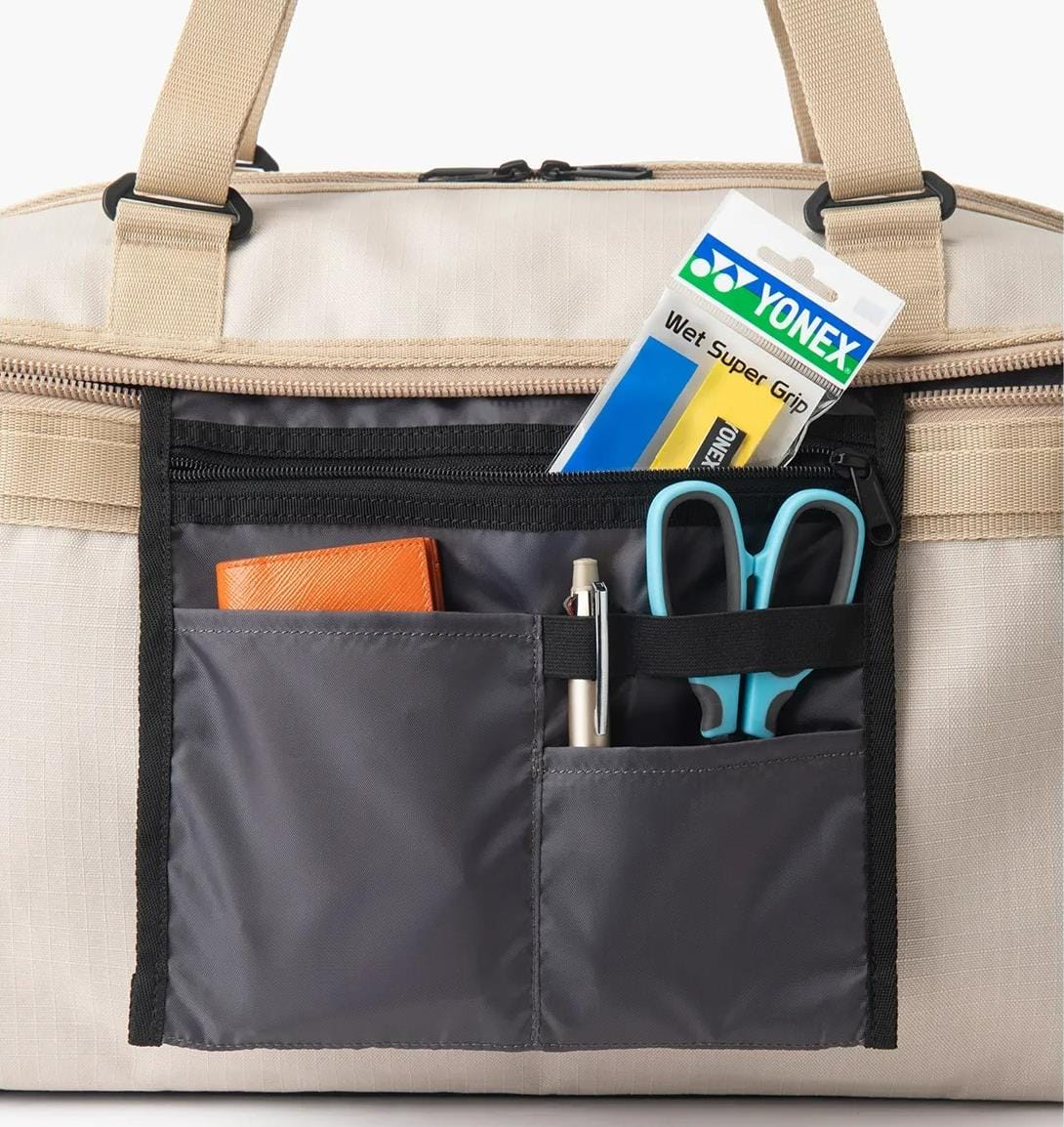 A close-up of the Yonex BA72611EX Gearlogic Tennis Boston Bag (Beige) front pocket—made from recycled polyester—shows a Yonex Wet Super Grip pack, brown wallet, pen, blue scissors, and black marker neatly arranged in compartments.