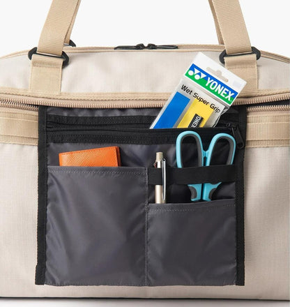 A close-up of the Yonex BA72611EX Gearlogic Tennis Boston Bag (Beige) front pocket—made from recycled polyester—shows a Yonex Wet Super Grip pack, brown wallet, pen, blue scissors, and black marker neatly arranged in compartments.