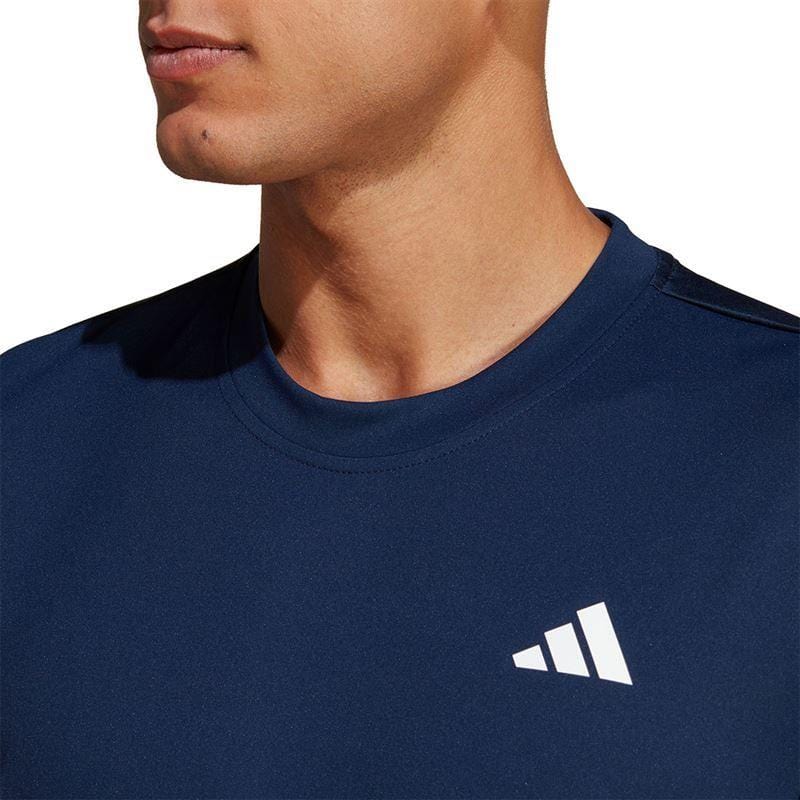 Close-up of a person wearing the ADIDAS Men's Club Tennis T-Shirt in navy blue, featuring a white abstract logo on the chest. The plain white background accentuates the AEROREADY shirt's sleek design, with no facial features visible to detract from its style.