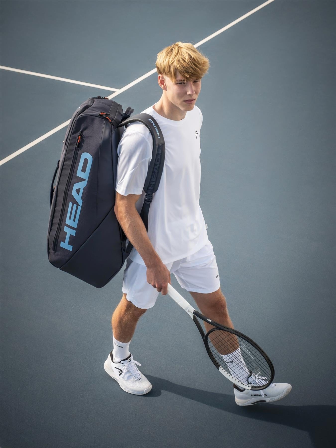 A young man in all-white athletic gear stands on a blue tennis court, holding a racket and carrying the HEAD Pro 12 Racket Tennis Bag XL - Navy by HEAD over his shoulder.