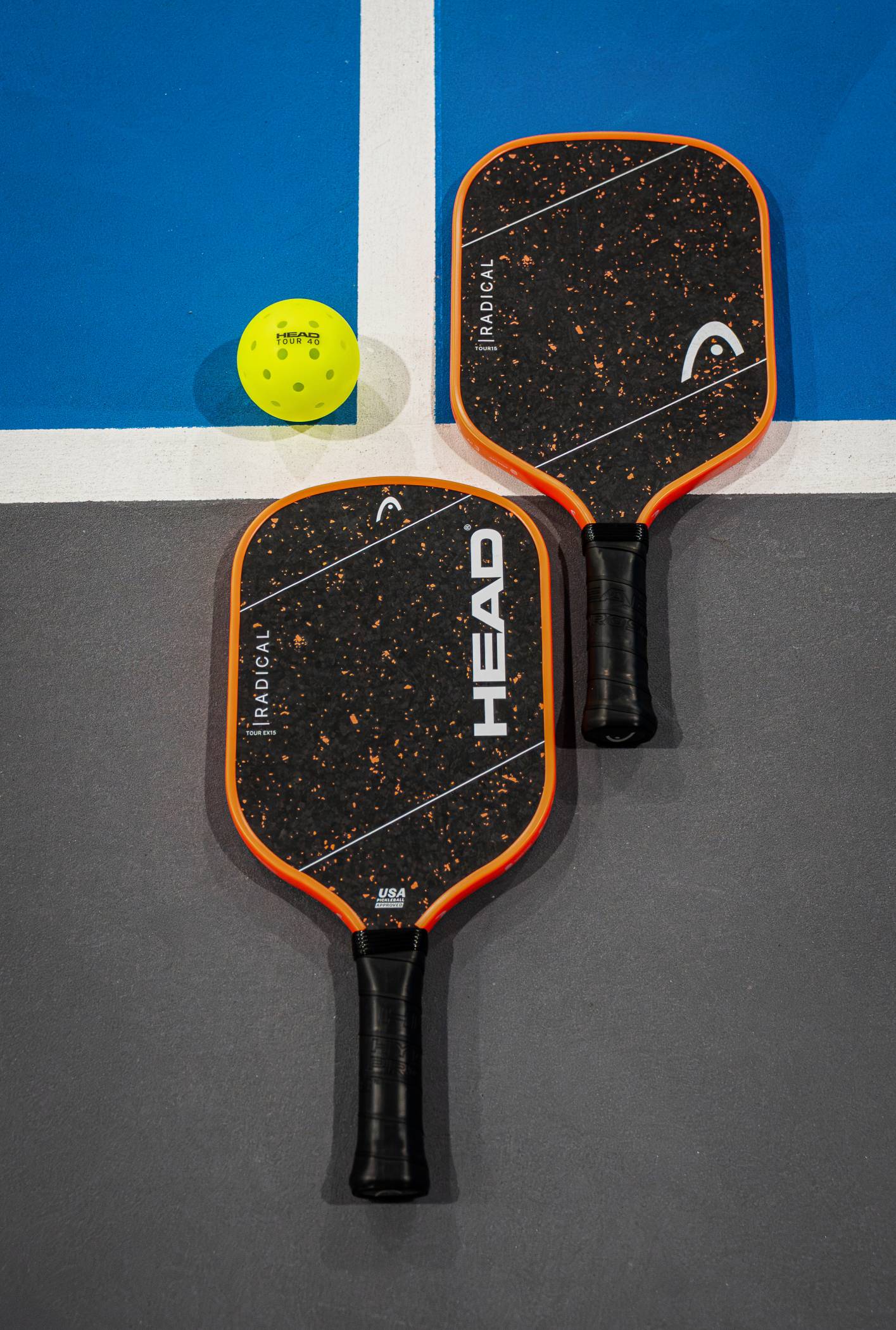 Two HEAD Radical Tour EX15 2026 Pickleball Paddles from HEAD, in striking black and orange with advanced thermoformed construction, rest side by side on the court beneath a yellow perforated pickleball, positioned near intersecting blue and white lines.