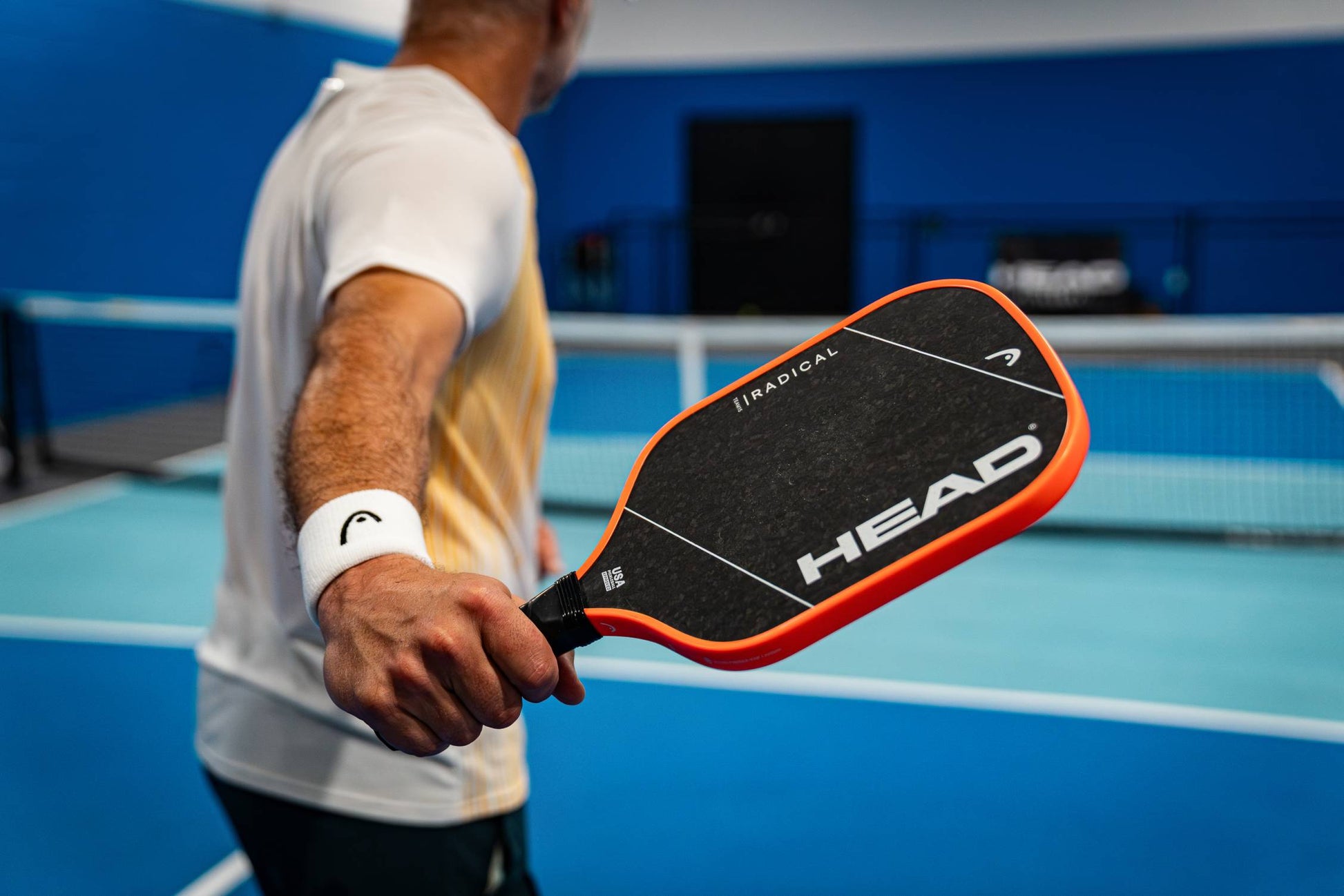 Dressed in sportswear, a player stands on an indoor pickleball court, gripping the HEAD Radical Team 15 2026 Pickleball Paddle by HEAD, featuring a TriFlex core. The spotlight is on the raw carbon paddle as the net and blue court background come into view.