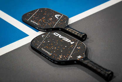 Two HEAD Radical Pro EX15 2026 pickleball paddles, featuring black raw carbon surfaces with speckled designs, lie crossed on a gray court near a blue and white boundary line.