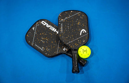 Two black HEAD Radical Pro 15 2026 pickleball paddles from HEAD overlap on a blue surface with a yellow perforated pickleball resting beside them. Equipped with the TriFlex Versatility Core and a raw carbon surface, these paddles deliver impressive power and control for every game.