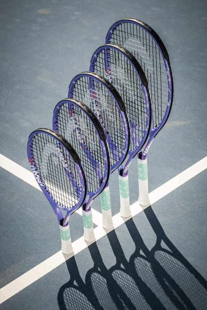 Five purple tennis rackets with white and green handles, including the HEAD Coco 17 2026 Junior Tennis Racket by HEAD, stand upright in a row on a blue tennis court, casting shadows near intersecting white lines—an ideal scene for anyone seeking the perfect beginner tennis racket for kids.