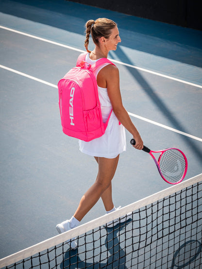 A woman in a white tennis outfit and sneakers smiles as she walks across a blue court, carrying the HEAD Base 17L Tennis Backpack in pink to match her racket, highlighting her stylish HEAD gear.