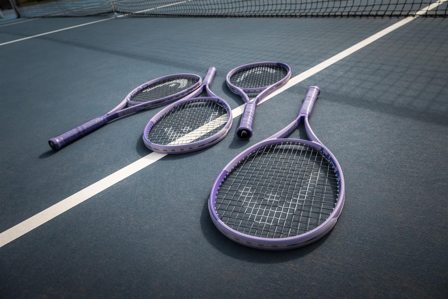 Four purple HEAD Boom MP L Alternate 2026 tennis rackets from HEAD rest on a blue court near the white boundary line, with the net in view.
